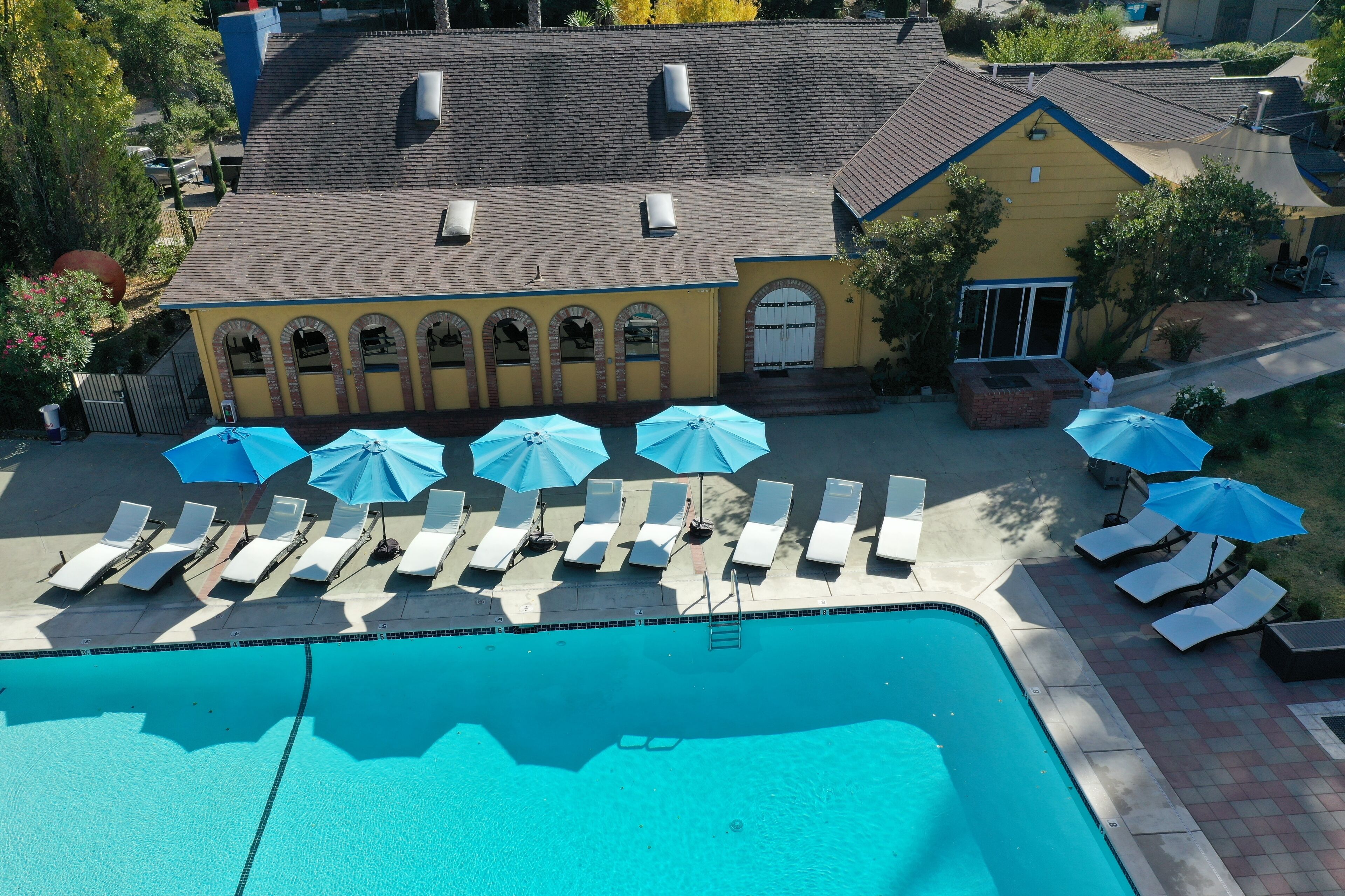 A swimming pool with sun loungers and umbrellas, a yellow building with a tiled roof, and a fenced yard.