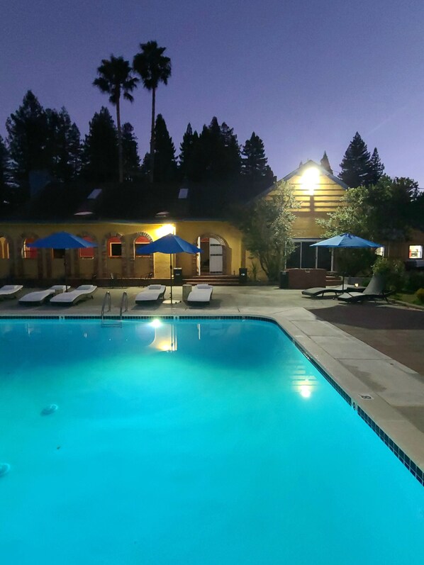 Una piscina con sillas de descanso y sombrillas, un edificio con ventanas de arco y palmeras al fondo.