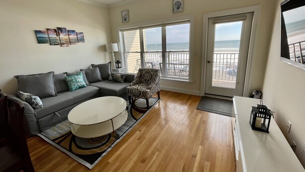 Ein Wohnzimmer mit grauem Sofa, einem gemusterten Sessel und einem runden Couchtisch. Große Fenster bieten Blick auf den Strand.