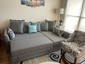Un sofá gris con varios cojines, un sillón estampado, una mesita con una lámpara y una alfombra con diseño de remolino.