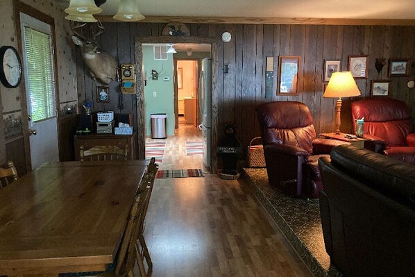 A wooden-paneled room with a dining table, two leather chairs, a lamp, and a deer head mounted on the wall.