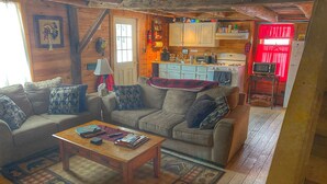 A cozy living room with a sofa, coffee table, and patterned rug. A kitchen with wooden cabinets and a microwave is visible in the background.