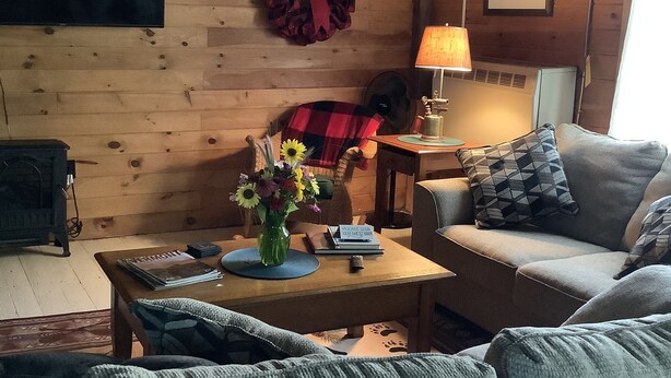 A cozy living room with a wooden wall, a fireplace, a sofa, a coffee table with a vase of flowers, and a sign for a guest book.