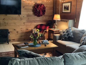 Un salon chaleureux avec un mur en bois, une cheminée, un canapé, une table basse ornée d’un vase de fleurs et un panneau indiquant le livre d’or.