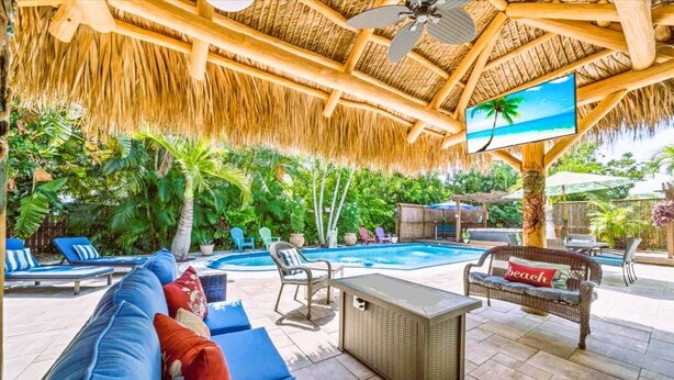 Un espace aménagé au bord de la piscine, doté d’un toit de chaume, de chaises longues, d’un canapé et d’un écran de télévision diffusant des images d’une plage.