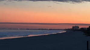 Une plage au coucher du soleil, avec un ciel dégagé, une mer calme et des bâtiments qui se dessinent à l’horizon.
