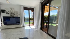 Un vaste salon doté d’un grand téléviseur à écran plat, d’un canapé confortable et d’une porte-fenêtre coulissante donnant sur une terrasse avec vue sur une piscine et un espace verdoyant.