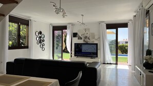 Un salon moderne avec un canapé en cuir noir, un téléviseur à écran plat fixé au mur et une grande fenêtre donnant sur un jardin.