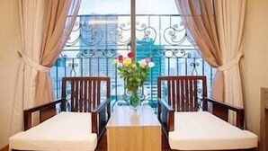 Ein Balkon mit zwei Holzstühlen und einem Tisch, einer Blumenvase und Blick auf ein Gebäude durch ein Glasgeländer.