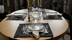 A round dining table set with plates, cutlery, and candles, positioned in a room with large windows and a view of trees and a building. - Modern Lux Cottage, Mtn Views, Close to Downtown (Bozeman)