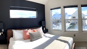 A bedroom with a large bed, two bedside tables, a dark headboard, and three windows providing a view of a snowy landscape. - Modern Lux Cottage, Mtn Views, Close to Downtown (Bozeman)