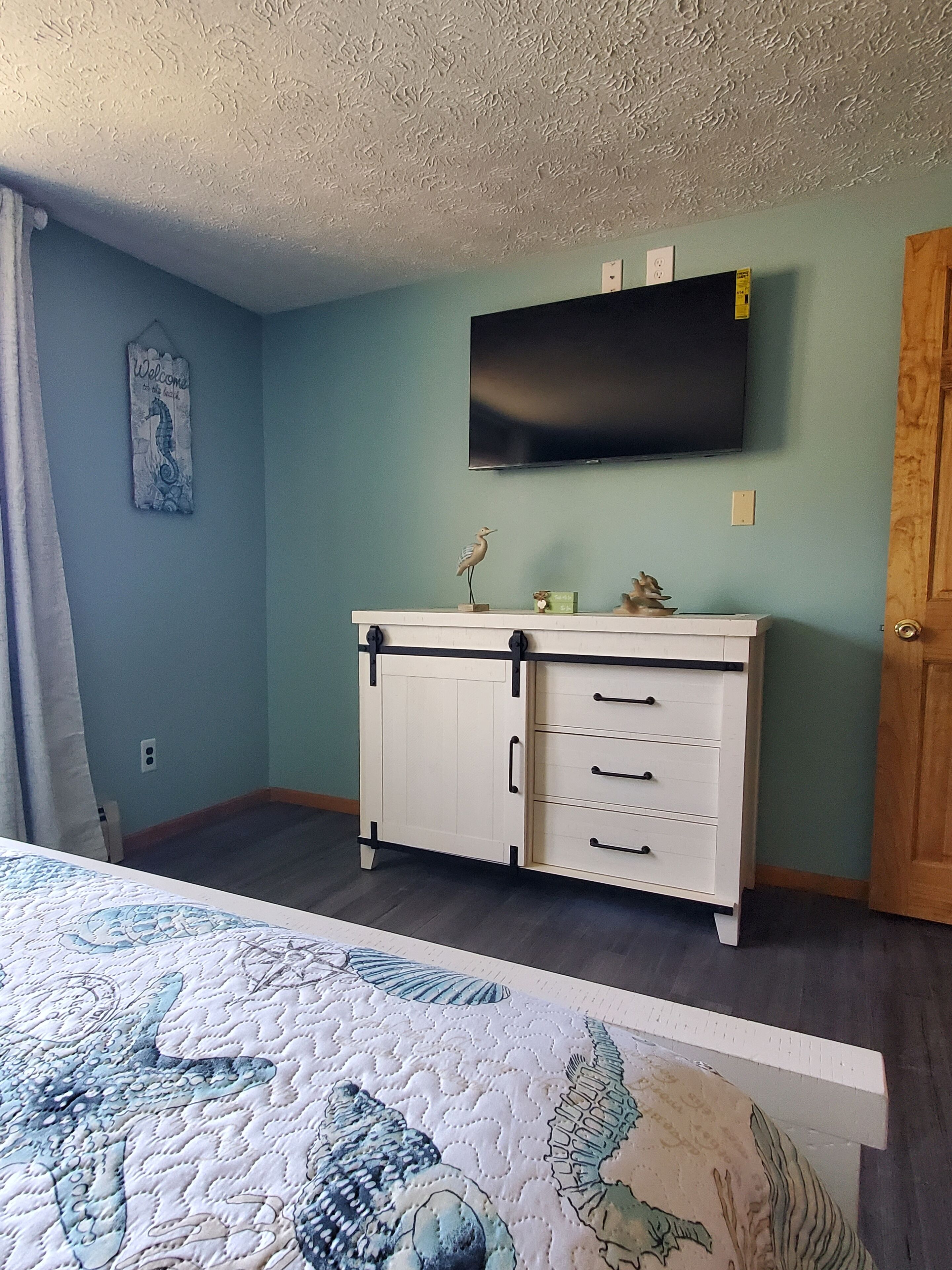 Une chambre à coucher avec un lit, une commode, un téléviseur fixé au mur et une porte.