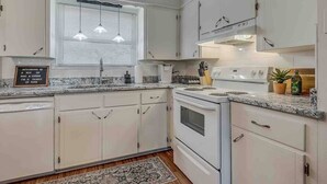 Una cocina con armarios blancos, horno empotrado, microondas y ventana con persianas.