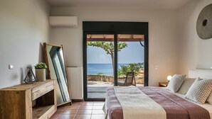 A bedroom with a bed, a wooden nightstand, a mirror, and a view of the sea through a sliding door. - Alekos Beach Houses-Eternity (Kefalonia)