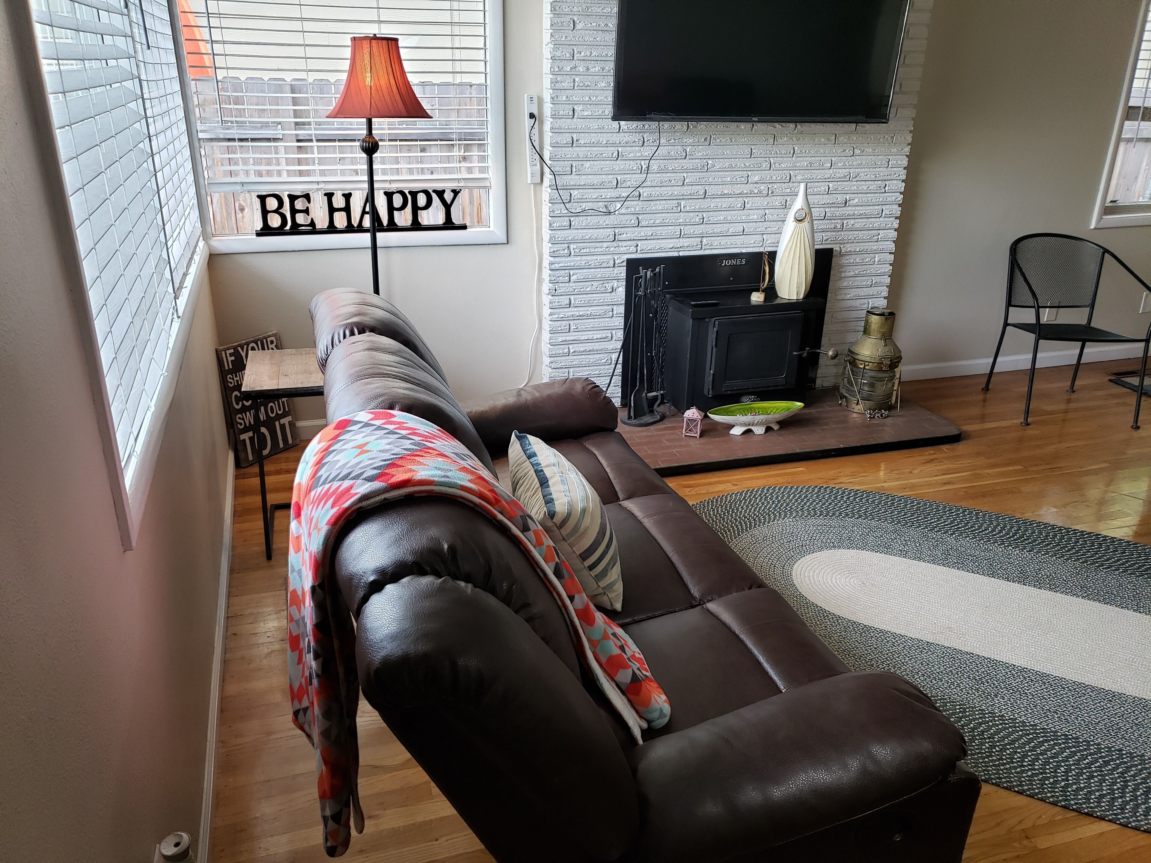 A living room with a brown leather sofa, a fireplace, a television mounted on the wall, and a window with blinds.