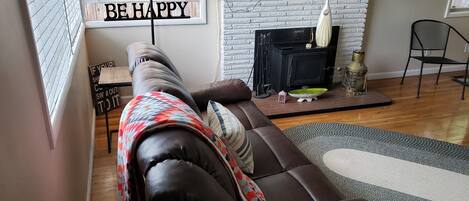 A living room with a brown leather sofa, a fireplace, a television mounted on the wall, and a window with blinds.