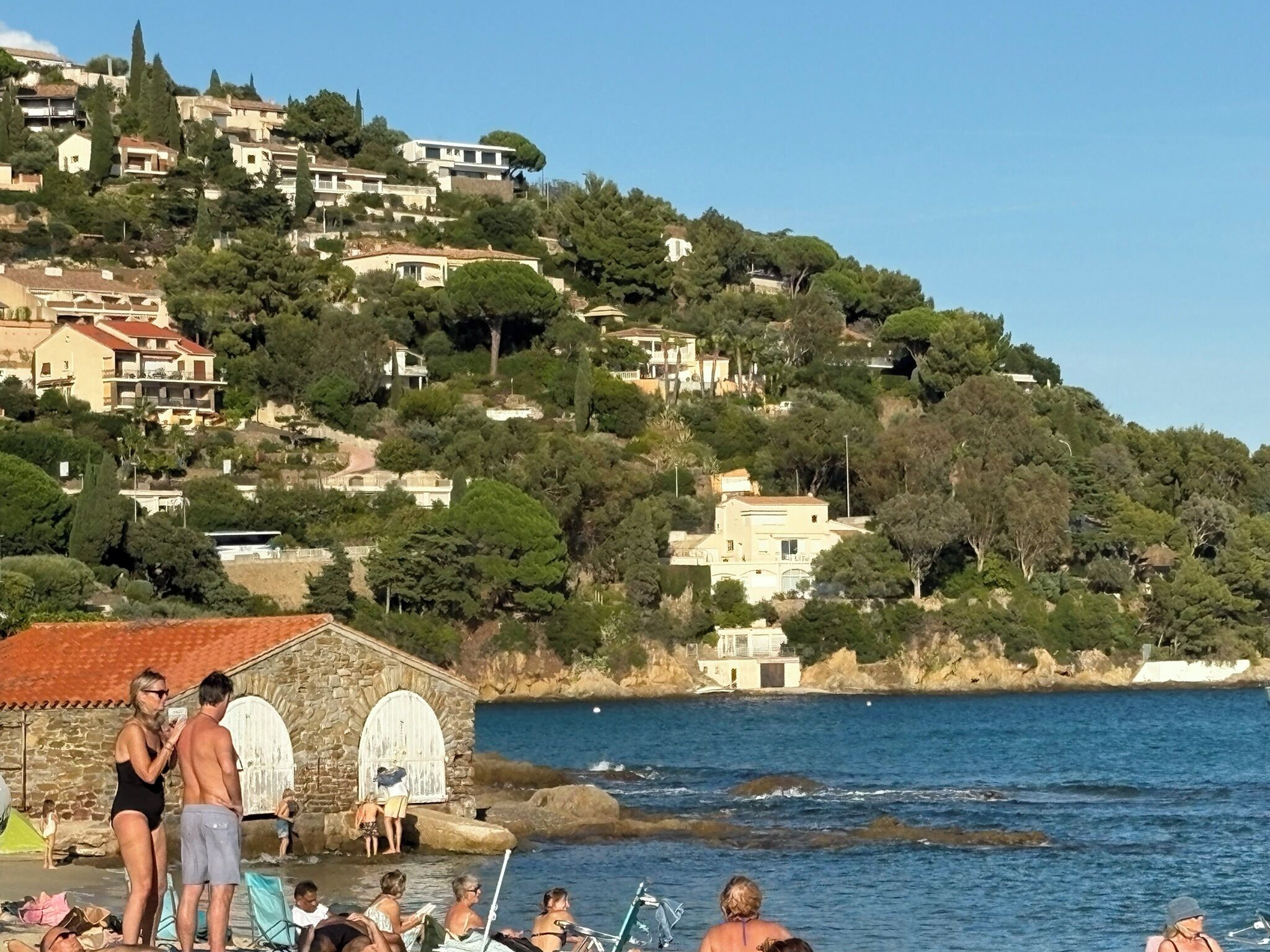 Une zone côtière avec des bâtiments construits sur une colline, une structure en pierre au bord de l’eau et des personnes près du rivage.