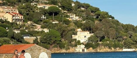 Une zone côtière avec des bâtiments construits sur une colline, une structure en pierre au bord de l’eau et des personnes près du rivage.