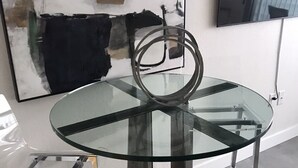 Ein moderner Essbereich mit Glastisch und Beinen aus Metall, ein zeitgenössisches abstraktes Gemälde an der Wand und Blick durch ein Fenster.