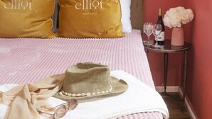Een netjes opgemaakt bed met een gestreept dekbedovertrek, twee kussens met de merknaam 'elliot', een klein nachtkastje met een fles wijn, glazen en een vaas met bloemen.