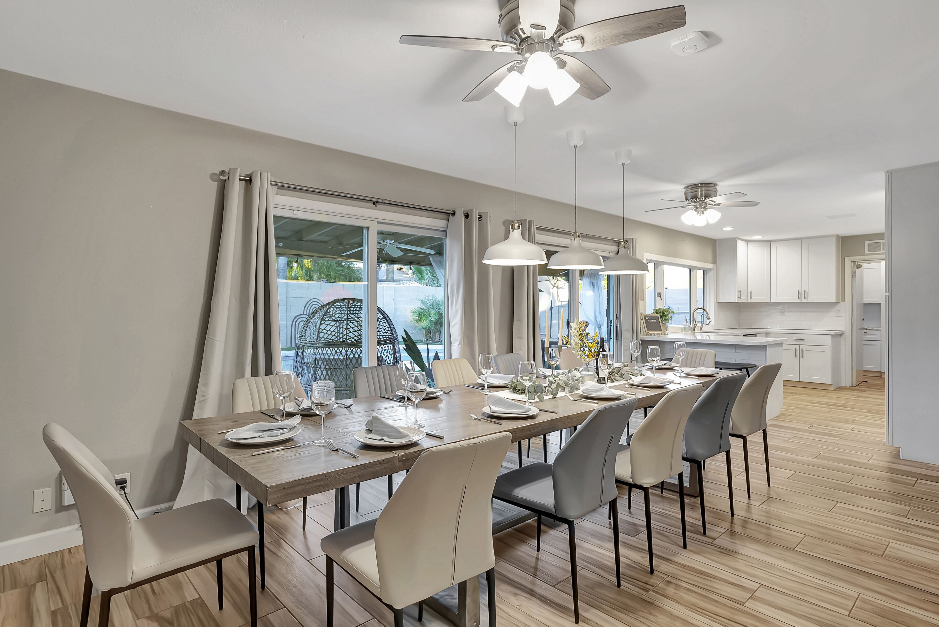 A dining area with a long table set for six, surrounded by chairs, with a ceiling fan above and a view of the kitchen in the background.