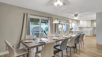 A dining area with a long table set for six, surrounded by chairs, with a ceiling fan above and a view of the kitchen in the background.