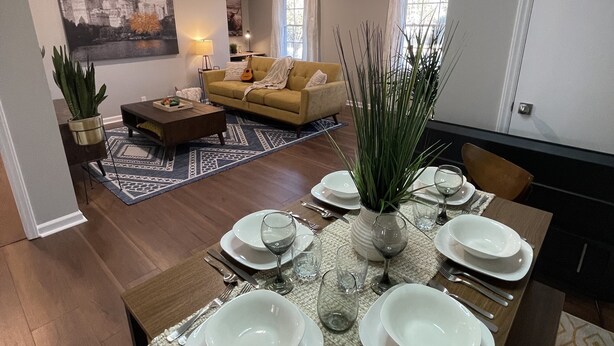 Ein Essbereich mit einem langen Tisch, gedeckt für sechs Personen, umgeben von einem Wohnzimmer mit einer Couch und einem Stadtbild.