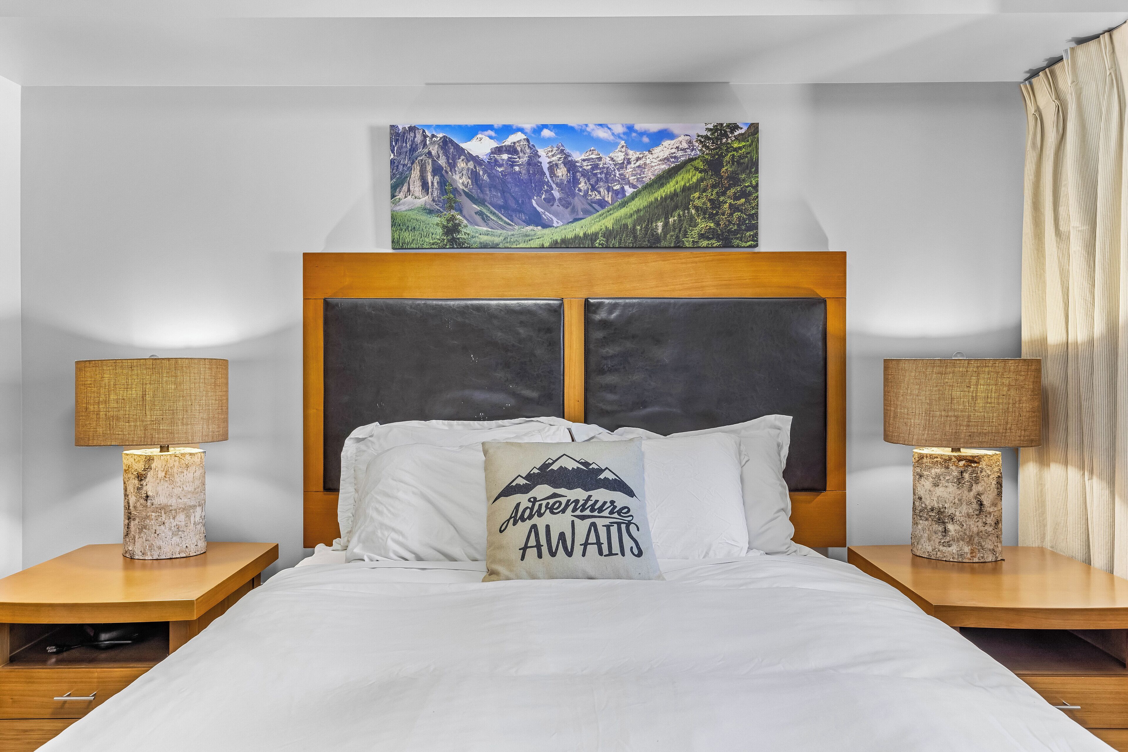 Ein Schlafzimmer mit einem hölzernen Kopfteil, einem Berglandschaftsbild über dem Bett, zwei Nachttischlampen und einem Kissen mit dem Aufdruck 'Adventure Awaits'.