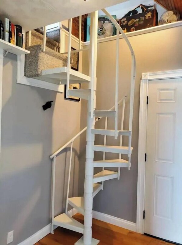 Una escalera espiral blanca con una zona de descanso alfombrada, una puerta blanca a la derecha y un estante con libros y adornos arriba.