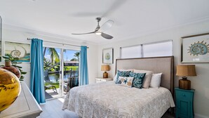 Ein Schlafzimmer mit einem großen Bett, einem Deckenventilator, einem Nachttisch mit Lampe und Blick auf einen Pool und Palmen durch eine Schiebetür.