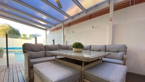 Un espace extérieur aménagé avec des meubles en osier et une table à plateau en verre, protégé par un auvent et offrant une vue sur une piscine.