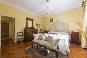 A bedroom with a bed, a dresser, a chair, a bench, a chandelier, and a mirror.