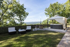 An outdoor patio with a hot tub, two white cushioned chairs, a covered seating area, and a stone fire feature.