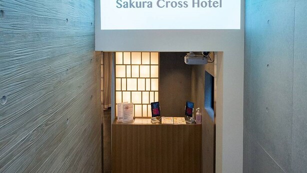Un área de recepción con un letrero que indica Hotel Sakura Cross, un mostrador de recepción y una escalera que sube a los pisos superiores.