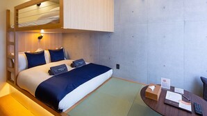 Habitación de hotel con literas, un escritorio con un control remoto y una mesita con una caja de pañuelos y un cartel.