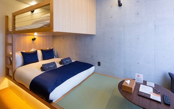Quarto de hotel com beliche, uma escrivaninha com controle remoto e uma pequena mesa com caixa de lenços e uma placa.