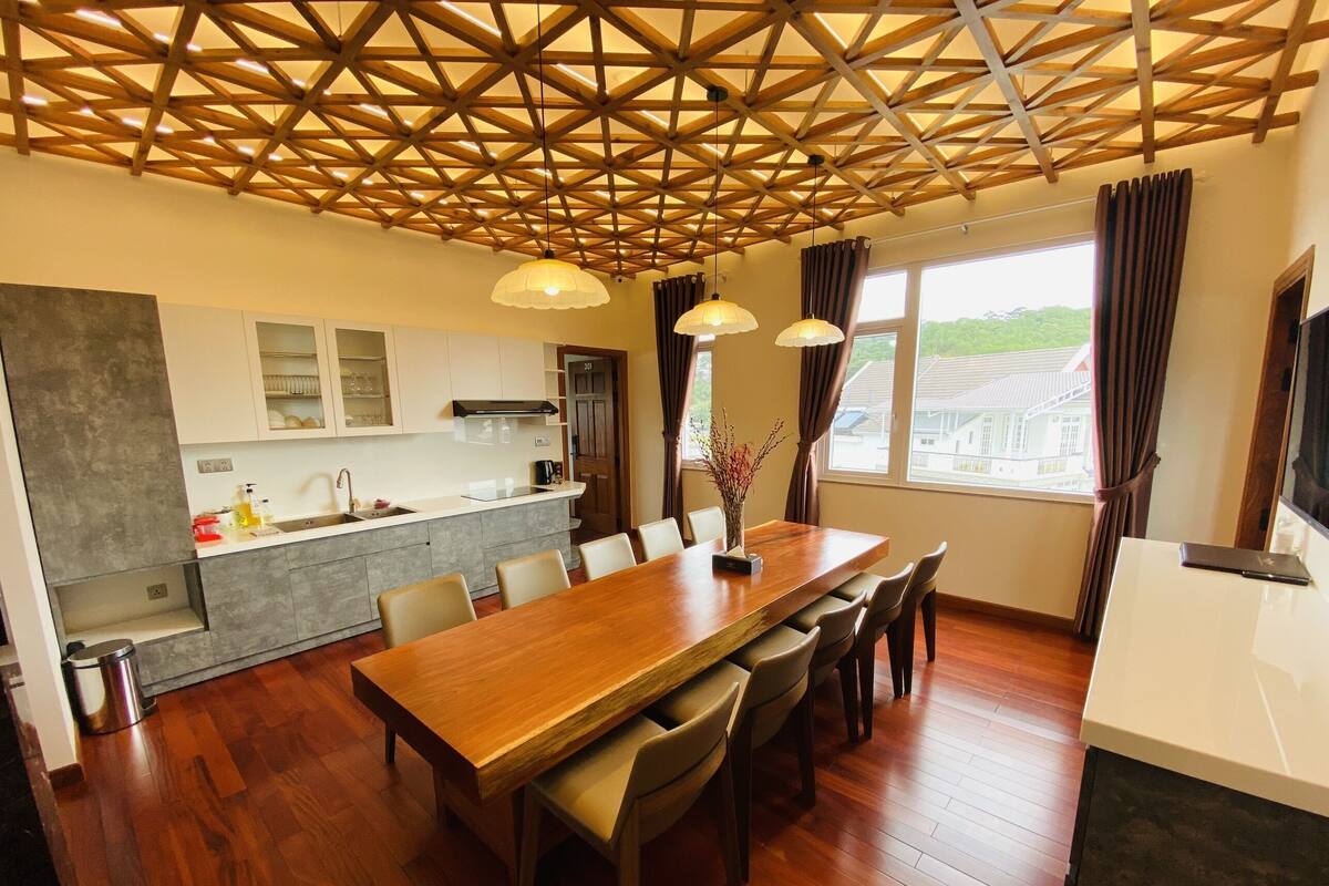 A modern kitchen with a wooden ceiling, a dining area with a large table and chairs, and a view of houses through the window.