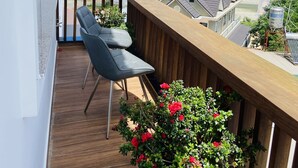 Un balcón con suelo de madera, dos sillas grises y una planta en maceta con flores rojas.