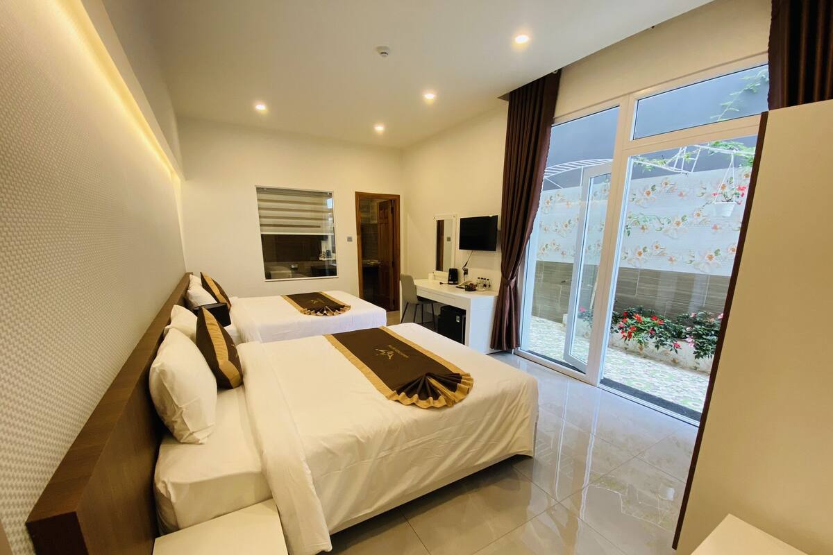 A hotel room with two beds, a desk, a TV, and a large window with a view of a garden.