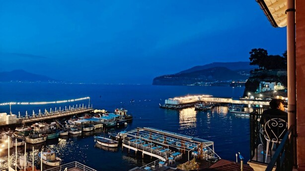 Un puerto deportivo al atardecer con barcos amarrados, un muelle que se adentra en el agua y una persona sentada en un balcón.