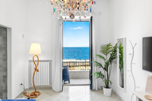A room with a view of the sea, a chandelier, a lamp, a potted plant, and a TV.
