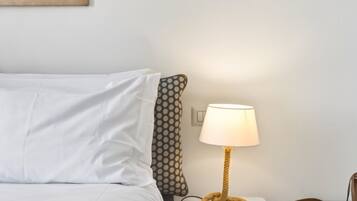 A neatly made bed with white bedding, a patterned pillow, a round table lamp with a rope base, and a brown leather bag on the side.