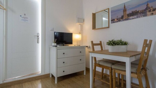 A room with a white door, a white dresser with drawers, a television on top, a table with chairs, a potted plant, and a framed picture on the wall.