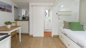 A compact room with a bed, a desk, a chair, a small table with a plant, a kitchen area, and a bathroom.
