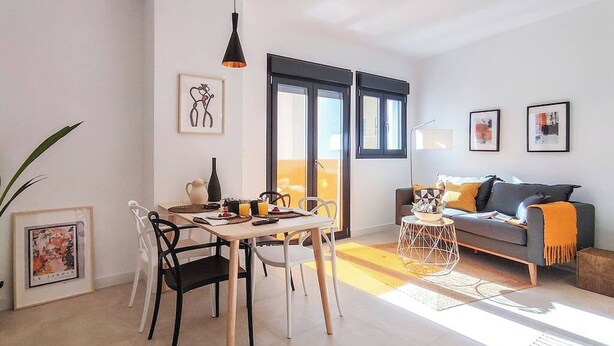 Una sala de estar moderna con un sofá, una mesa de comedor con sillas y un balcón con ventanales amplios.