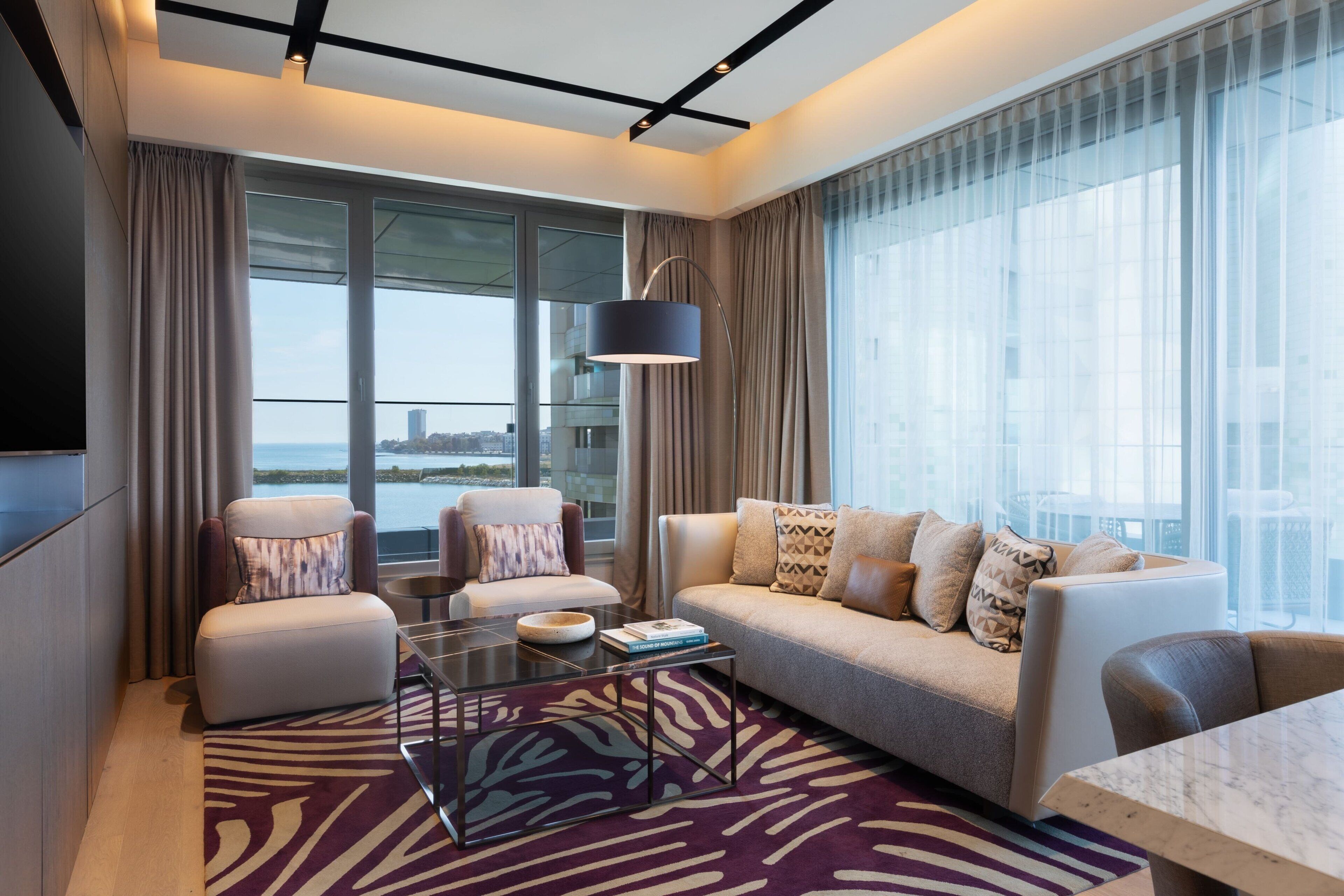 Una habitación de hotel moderna con un sofá, sillones, una mesa de centro de vidrio y vistas a la ciudad.