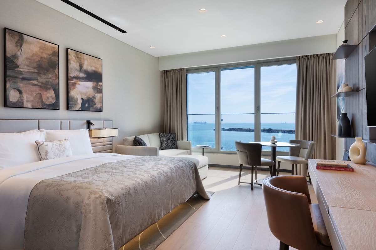 Habitación de hotel moderna con una cama grande, una zona de estar con mesa y sillas, y un amplio ventanal con vistas al mar.