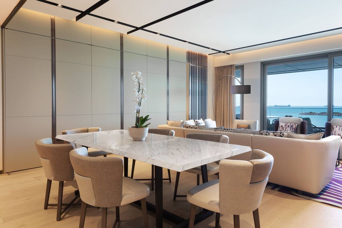 Una sala de estar moderna con una mesa de comedor de mármol, sofás beige y un ventanal con vistas al mar.