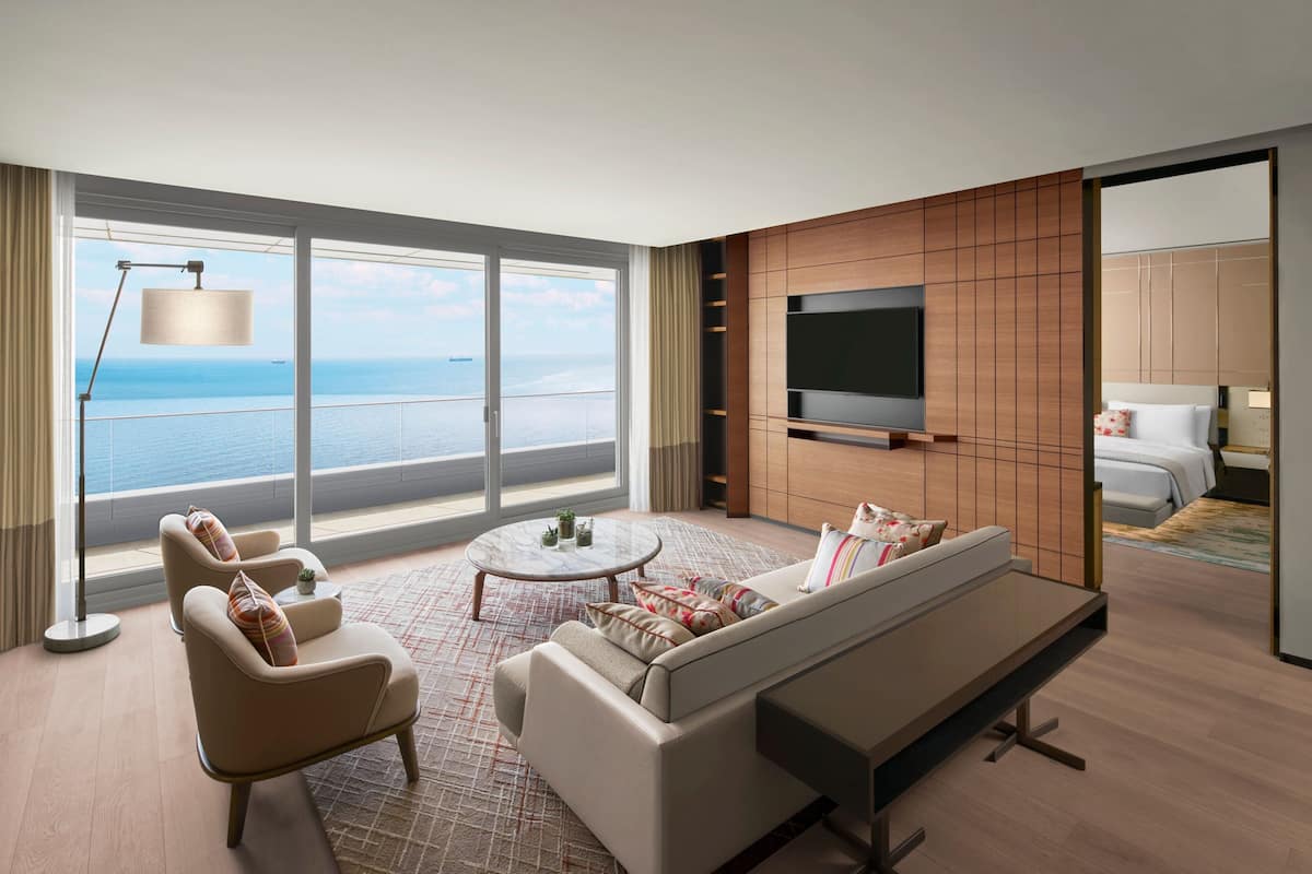 Habitación de hotel moderna con un amplio ventanal que ofrece vista al mar, un sofá, sillones, una mesa de centro y un televisor de pantalla plana montado en la pared.