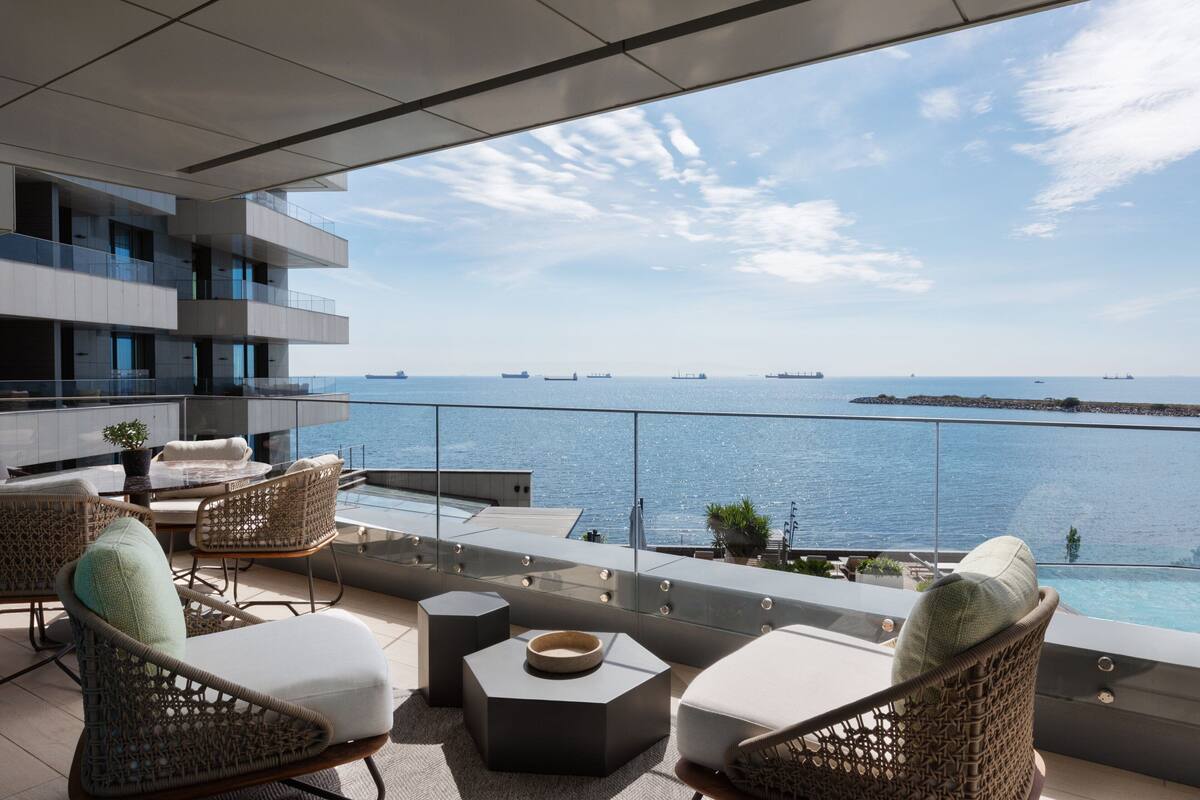 Un balcón moderno con mobiliario de mimbre, barandilla de vidrio y vista al mar.